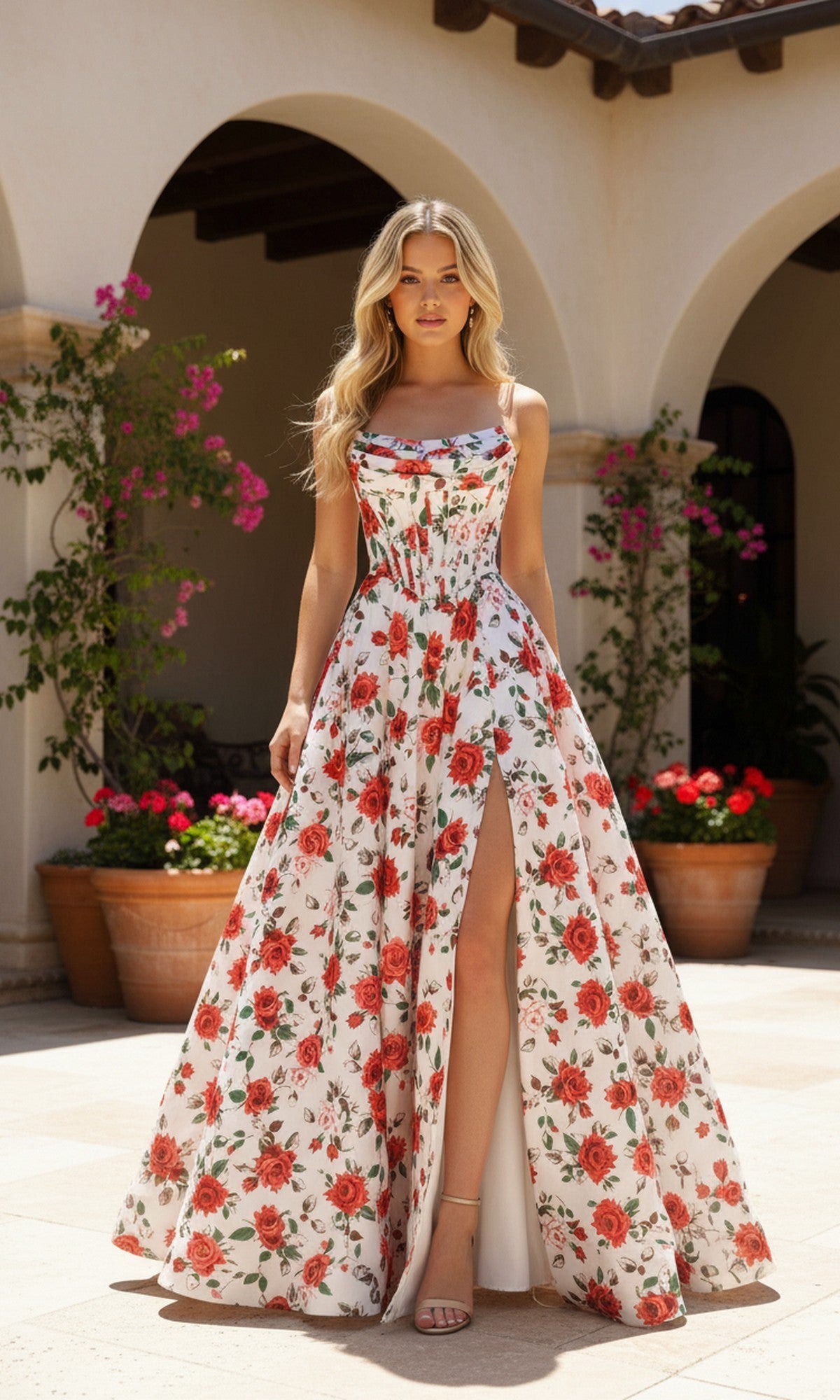 A woman wearing the PromGirl Exclusive 250229 long prom dress in ivory/red print with a sleeveless design and high slit stands outdoors near potted plants and archways.